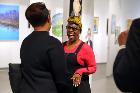 a woman wearing a headscarf and glasses laughs while talking to someone else in an art gallery