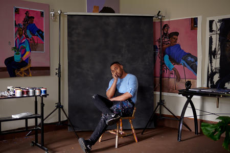 photo of a Black man sitting in an artist studio with portraits of himself on the walls