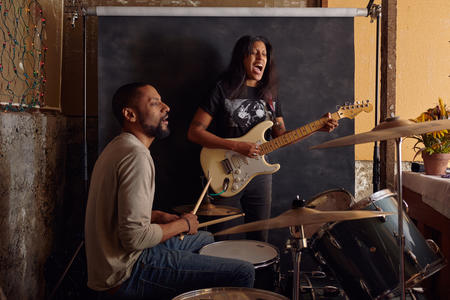 Two people in front of a black screen, one is on drums, the other sings behind a guitar