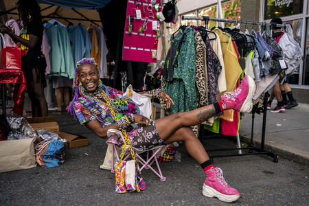 Person sits in a chair and stretches their leg. their shoes are pink and they are wearing colorful clothes.