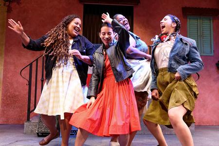 Four women in skirts are dancing in a group, having fun. They are on a stage with a (muted) red backdrop that looks like the façade of a house with iron staircase.
