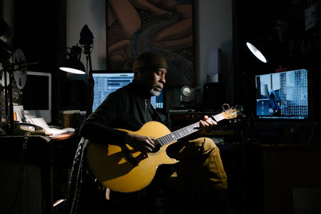 Person in studio with guitar lit by a light and blue screens behind 