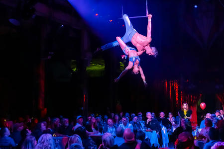 Two people hang from a trapeze, lit by a spotlight and with a crowd below