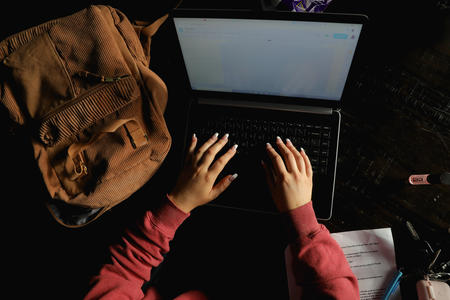 Two hands extend over a laptop keyboard see from above next to a brown backpack.