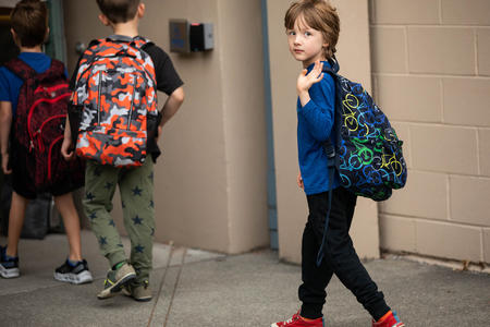 A young student wearing a backpack turns to wave while walking through a school door.