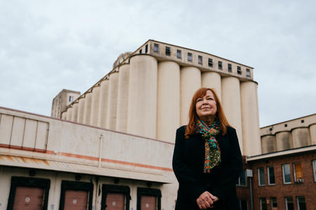 Person dressed in dark clothing wearing a colorful scarf and with red hair stands in front of a large, beige building featuring grain silos