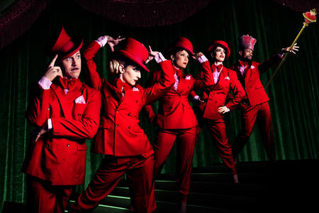 Six figures dressed in bright red clothing in a dark room, stand on stairs ascending from left to right