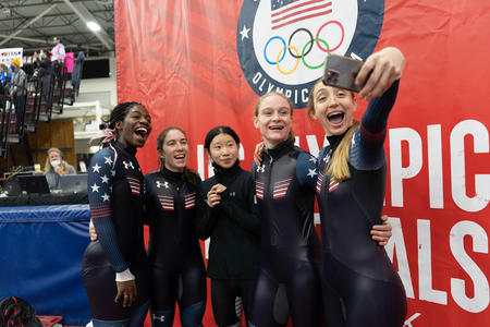 Five athletes in dark suits taking a selfie together in front of a red backdrop