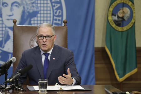 Jay Inslee sits at a table with the Washington state seal and flag behind him.