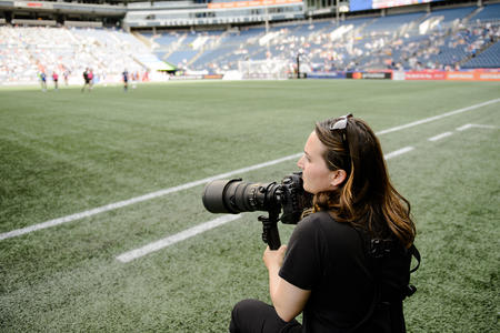 A woman holding a camera while standing on the sidelines of a soccer pitch
