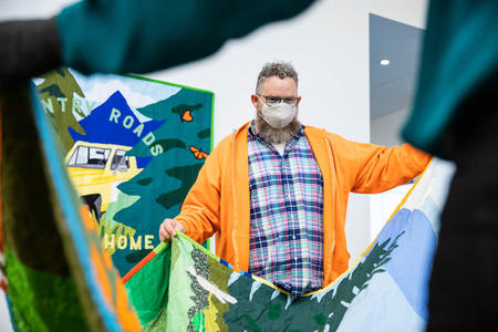 Person in plaid shirt with orange sweater, wearing a mask over his beard, holding a quilt and standing in front of a quilt