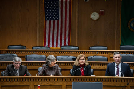 Four people in business attire sitting at a wooden desk in front of an American flag