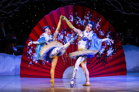 Two people in blue and white costumes in front of a red backdrop dance like ballerinas, holding each other's hands
