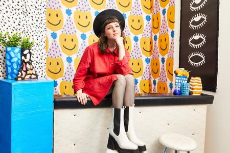 White woman in red shirt dress and white and black boots, wearing a black beret, sits on a ledge. next to her stands a blue column with colorful vases, the wallpaper bears a pattern of yellow smiley faces