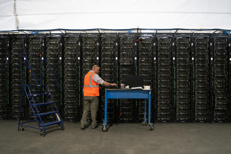 A worker with a rolling cart stands in front of a tall row of computer servers inside a warehouse