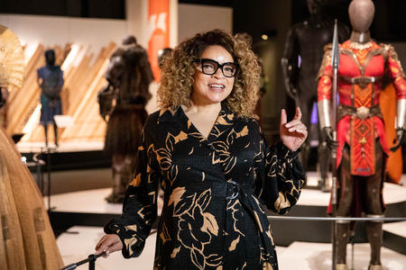 a woman with curly hair and glasses stands in front of museum installation of a futuristic film costume