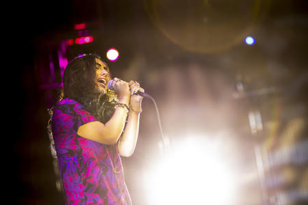 Person on left side of photo facing the right side of the photo, light behind them shining into camera. The person is singing into a microphone. The singer has long dark hair.