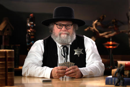 Knute Berger is seen in a western hat wearing a Marshal's star and holding a hand of cards.