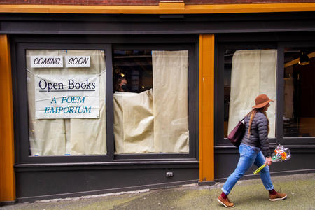 Person in orange hat and flowers in hand walks by window that is still covered in paper. Behind the window, a person is taking the paper off. To her left, a sign reads: Open Books: A Poem Emporium