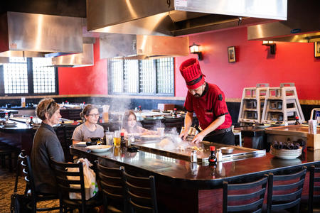 A chef in a tall red hat and shirt grills up Japanese food on a hot stove in front a group of three people. 