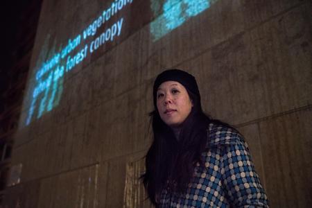 a woman with long dark hair wears a beanie and stands below words projected on a wall in blue light