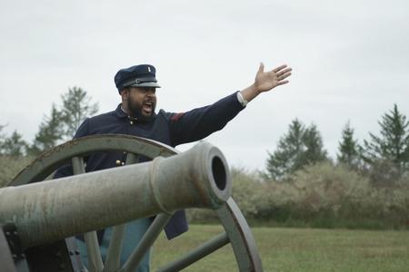 A movie still featuring a Black man in military dress shouting next to a cannon