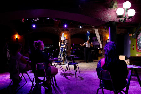 stage lit with purple light, some people on chairs, some empty chairs. in the white spotlight is an actor wearing a hat that looks like a building