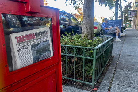 A red newspaper box with copies of the Tacoma Weekly, with a man sitting on a bench in the background