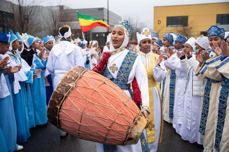 A woman plays a drum during a celebration outside with people wearing traditional white garb