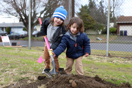 two smiling children dig a hole in a field lined with chainlink fence