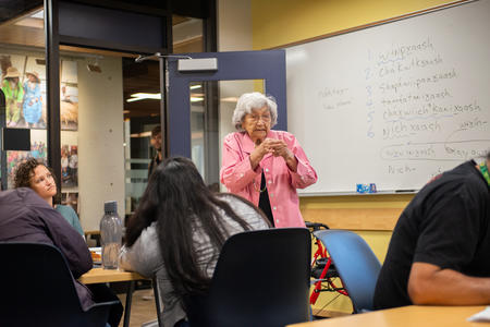 An elderly woman stands at the front of a classroom speaking to students