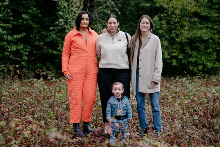 Three people stand amid various plants and trees in the background. The person on the left wears a bright orange jumpsuit, the person in the middle a beige fleece, and the person on the right a beige jacket and jeans. A small kid is standing in front of them, in the middle.