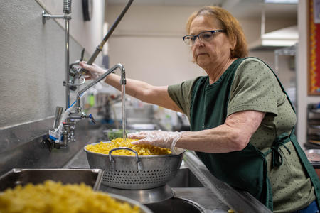 A school kitchen employee rinses curly pasta in a sink.