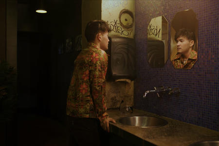 A wide shot of an Asian man staring at his reflection in the mirror pensively