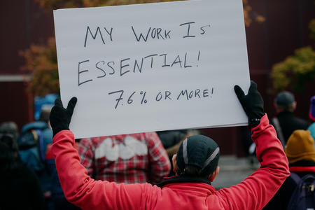 a person holds a sign at a rally outside city hall reading "my work is essential" 