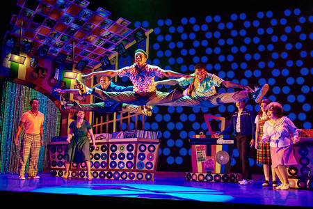On a colorful stage set, people in brightly patterned outfits do splits in the air as onlookers watch; the lighting is bright hues of purple, blue and pink
