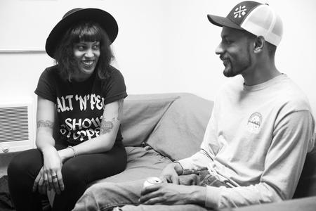 a black and white photo of a woman and a man on a couch, smiling at each other, both wearing hats