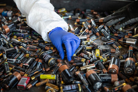 a gloved hand holds batteries over a pile of many batteries