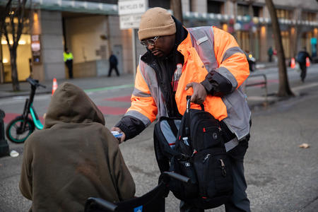 a man in an orange safety jacket hands out socks to a person on the street in downtown seattle