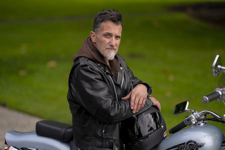A man wears a leather jacket and sits on a motorcycle. He looks at the camera with a serious expression.