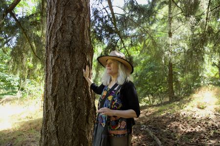a woman stands next to a tree, her hand placed on its trunk, as she looks away from the camera