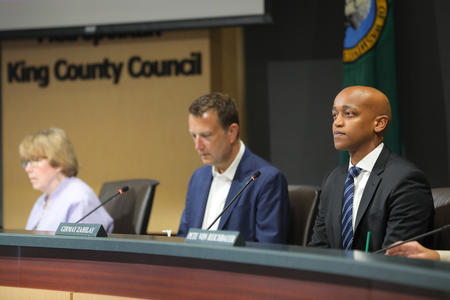 Three people sit at a dais that is in front of a sign that says "King County Council."