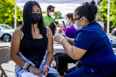 A seated health care professional holds a needle and a bandage while a seated middle school student looks away.