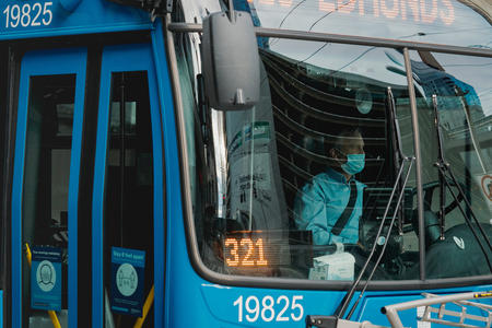 A driver with a mask is seen through the window of a blue bus.