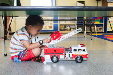 A child sits under a table and plays with a toy firetruck.