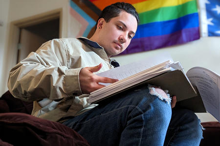 A person sits while reading documents, an LGBTQ+ pride flag is on the wall in the background.