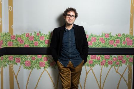 photo of a man with brown hair and glasses standing against a wall painted with flowers