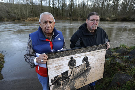 Two people on a riverbank hold a historical photo of people fishing in that river.