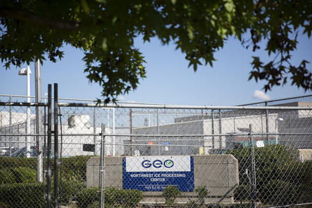 A sign behind a fence says "GEO Northwest ICE Processing Center." White buildings are in the background.