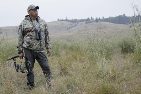 Man standing wearing camo in knee high grass holding a bow and looking over horizon